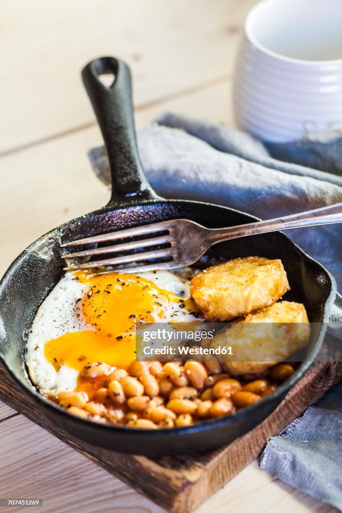 Fried egg, baked beans and hash browns in frying pan on wooden board