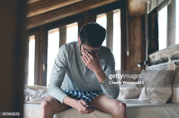 tired young man with smartphone sitting on bed at home squinting - waking up in bed stock pictures, royalty-free photos & images