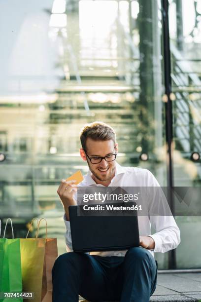 smiling businessman holding credit card and using laptop next to paper bags - mobile banking stock-fotos und bilder