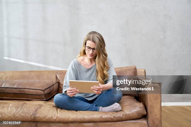 blond woman sitting on leather couch using tablet - assis en tailleur photos et images de collection
