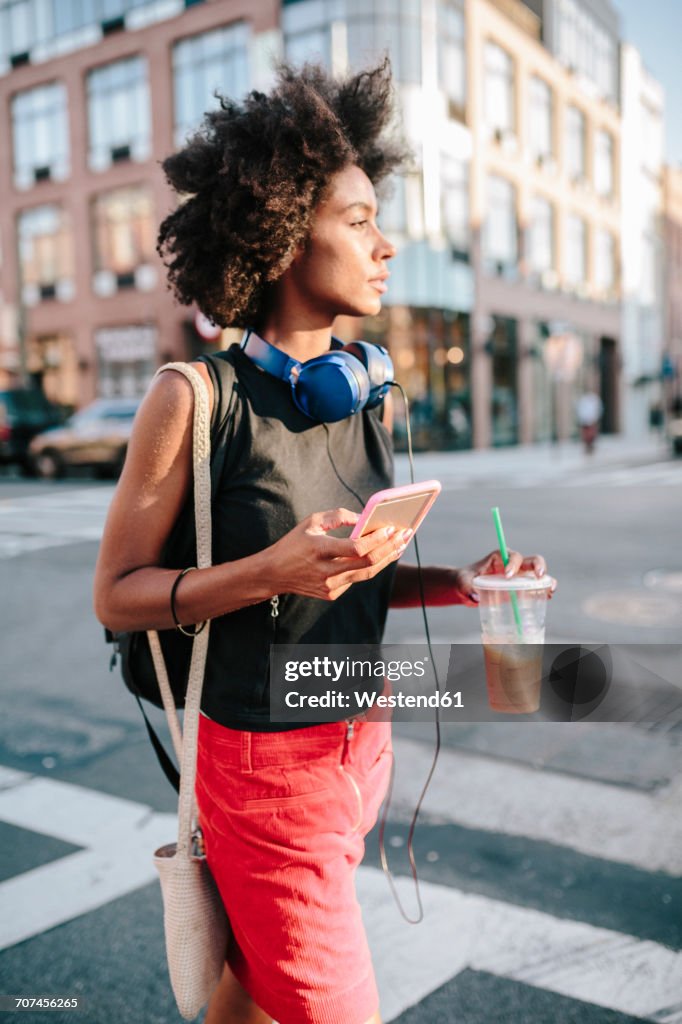 Young woman with headphones and smart phone crossong street in Brooklyn, carying take away drink