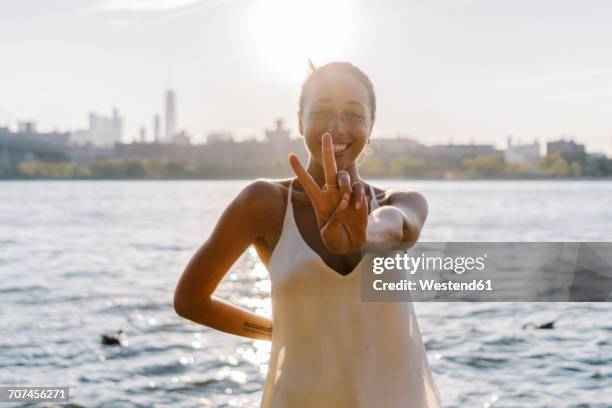 young woman in brooklyn standing at east river giving victory sign - victory sign stock-fotos und bilder