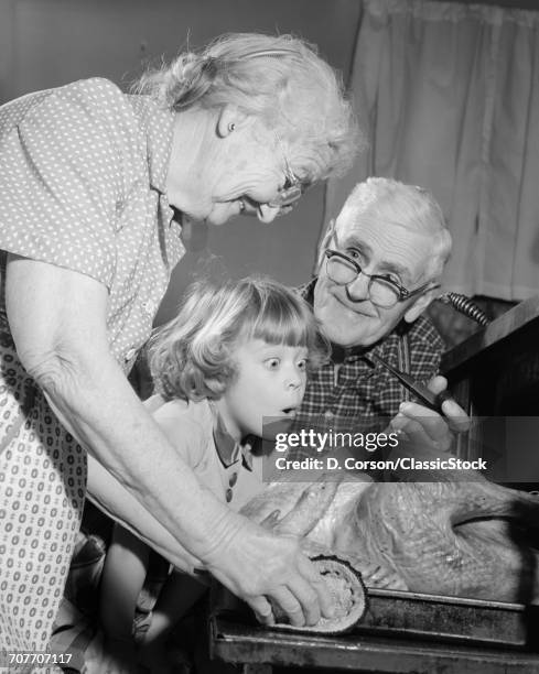 1950s 1960s EXCITED LITTLE GIRL GRANDDAUGHTER WITH GRANDPARENTS WATCHING ROAST TURKEY COMING OUT OF OVEN