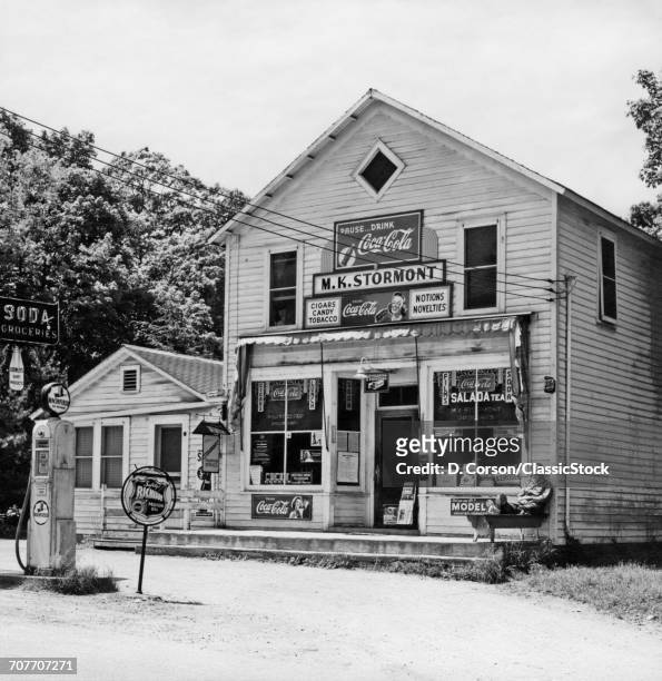 1940s 1950s MAN STILLING ON PORCH GENERAL STORE AND RICHFIELD GAS STATION AMID SIGNS FOR COCA-COLA SALADA TEA PUBLIC TELEPHONE