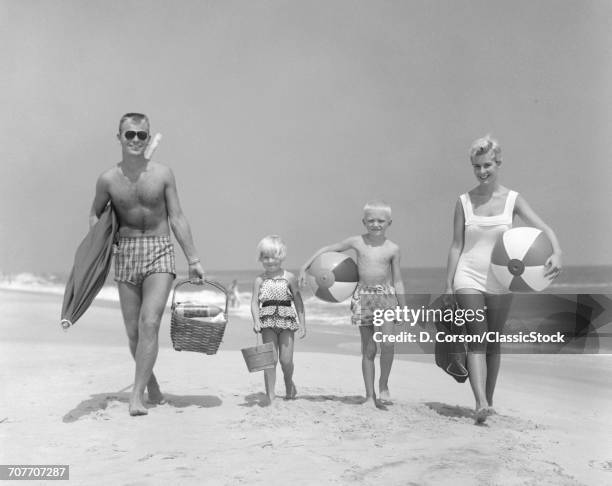 1950s FAMILY OF FOUR WALKING TOWARDS CAMERA WITH BEACH BALLS UMBRELLA PICNIC BASKET AND SAND BUCKET