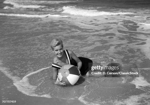 1960s SMILING WOMAN IN BATHING SUIT LAYING IN THE OCEAN SURF HOLDING A BEACH BALL LOOKING AT CAMERA