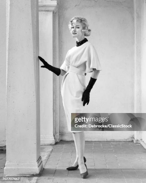 1960s FULL LENGTH PORTRAIT ELEGANT BLONDE WOMAN WEARING DRESS WITH FULL DRAPED SLEEVES LONG GLOVES POSING BY COLUMN OUTDOORS