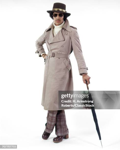 1970s AFRICAN AMERICAN MAN WEARING TRENCH COAT AND HAT LEANING ON UMBRELLA LOOKING AT CAMERA