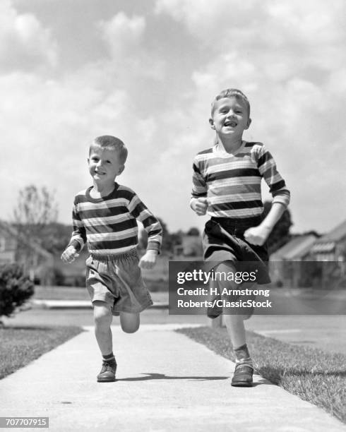 1950s TWO SMILING BOYS BROTHERS WEARING STRIPED SHIRTS SHORTS RUNNING ON SIDEWALK LOOKING AT CAMERA SUBURBAN NEIGHBORHOOD