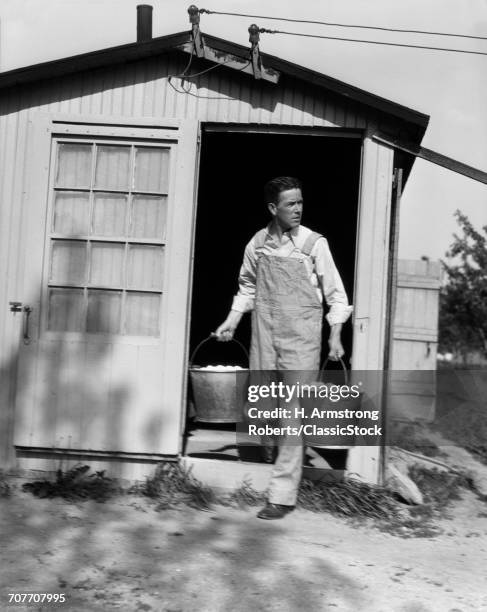 1920s 1930s MAN IN BIB OVERALLS COMING OUT OF A FARM BUILDING WITH TWO PAILS FULL OF CHICKEN EGGS