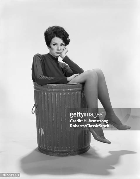 1960s UNHAPPY SAD DEPRESSED WOMAN IN TRASH CAN WEARING HIGH HEELS LOOKING AT CAMERA