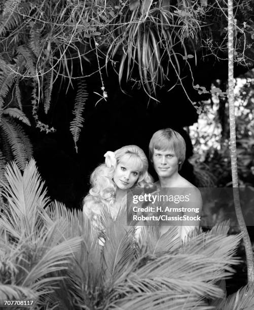 1970s BLONDE COUPLE POSED AS ADAM AND EVE LOOKING AT CAMERA STANDING NAKED BEHIND TROPICAL FOLIAGE EVE HOLDING APPLE