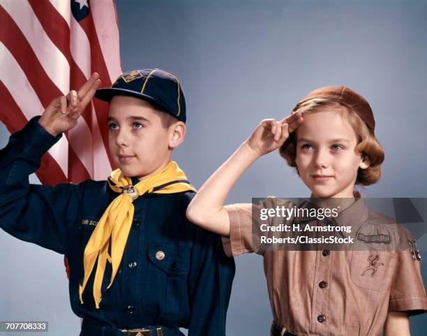 1960s BOY AND GIRL IN CUB SCOUT AND BROWNIE UNIFORMS STANDING BY AMERICAN FLAG SALUTING