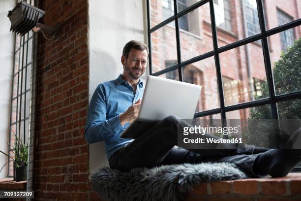 mature man sitting on window sill, using laptop - apartamento tipo loft fotografías e imágenes de stock