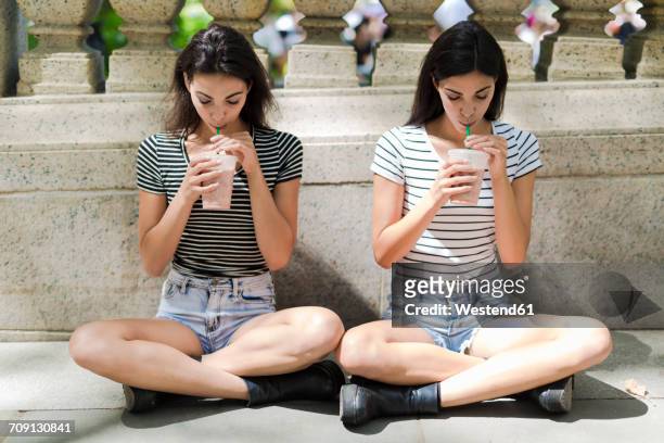two twin sisters sitting outdoors having a takeaway drink - hot pants stock-fotos und bilder