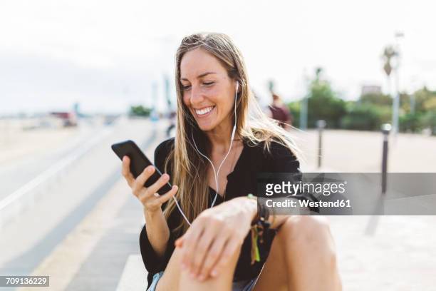 smiling young woman with cell phone and earbuds on waterfront promenade - differential focus fotografías e imágenes de stock