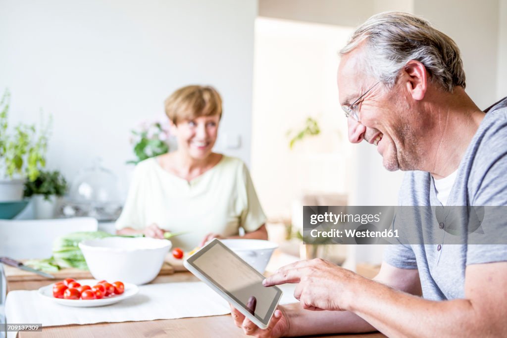 Senior man at home using digital tablet with woman preparing salad in background