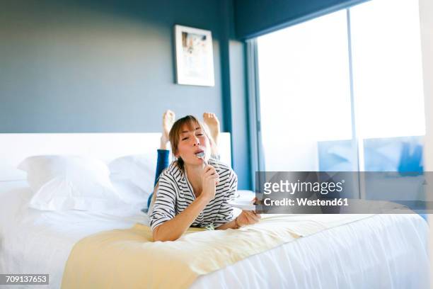 young woman eating piece of vegan cake, lying on bed, licking spoon - indulgence photos et images de collection