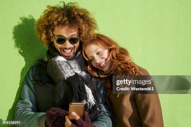 happy young couple looking at cell phone at green wall - winter woman phone stock-fotos und bilder