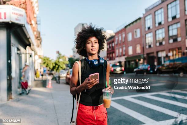 young woman with headphones and smart phone crossong street in brooklyn, carying take away drink - brooklyn new york stock-fotos und bilder