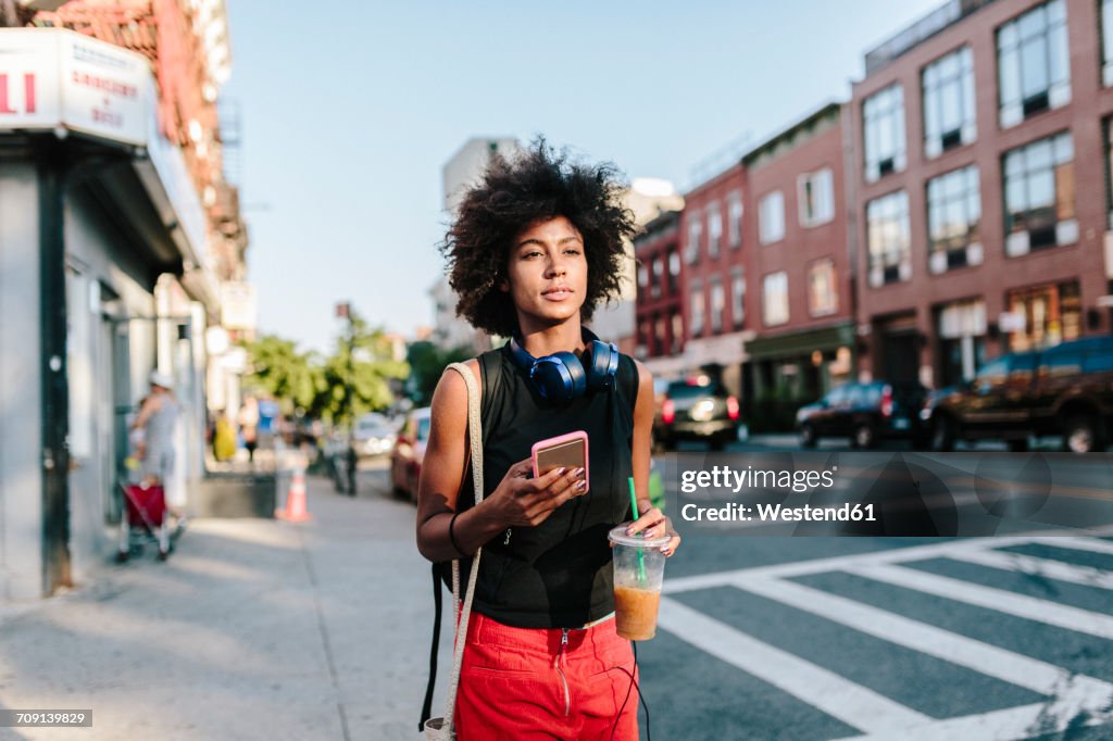 Young woman with headphones and smart phone crossong street in Brooklyn, carying take away drink