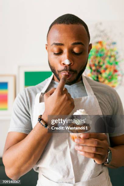 young man eating a cup cake with whipped cream, licking finger - macchie di colore foto e immagini stock