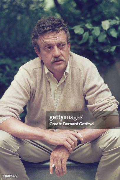 American novelist and humanist Kurt Vonnegut wears a cardigan sweater as he sits on a step and smokes a cigarette, probably a Pall Mall, early 1970s.