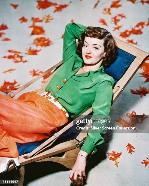 American actress Bette Davis , relaxing in a deckchair surrounded by autumn leaves, circa 1940.