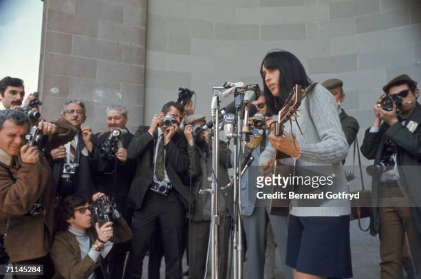 Press photographers take pictures of American folk singer and social activist Joan Baez as she sings and plays guitar into NBC microphones on stage...