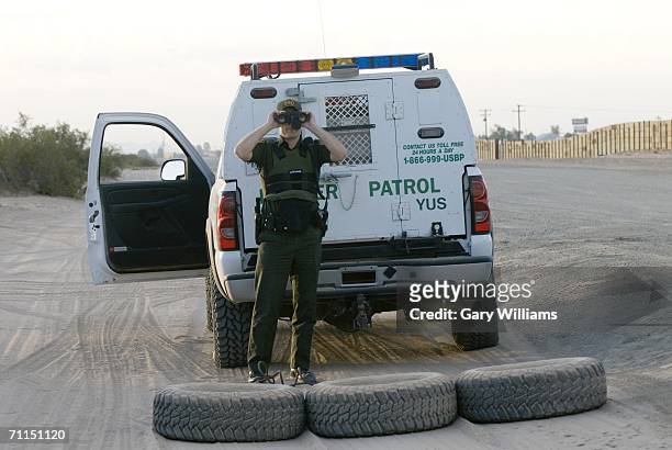 Border Patrol agent uses a pair of binoculars as he looks at an apparent illegal immigrant try to climb over a 15 foot high border fence which leads...
