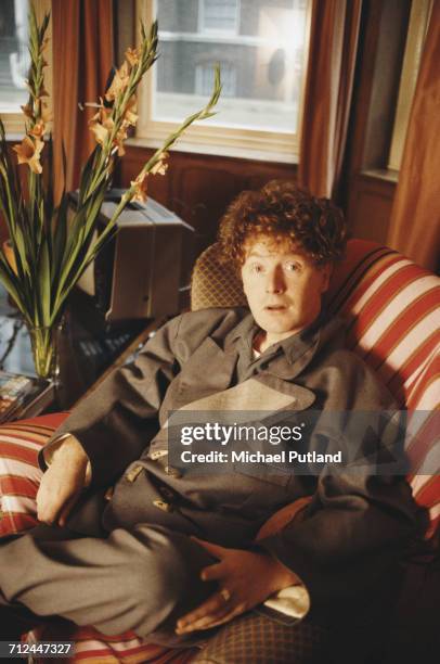 English performer and manager, Malcolm McLaren pictured seated in a chair wearing a double breasted jacket at home in London in 1983.