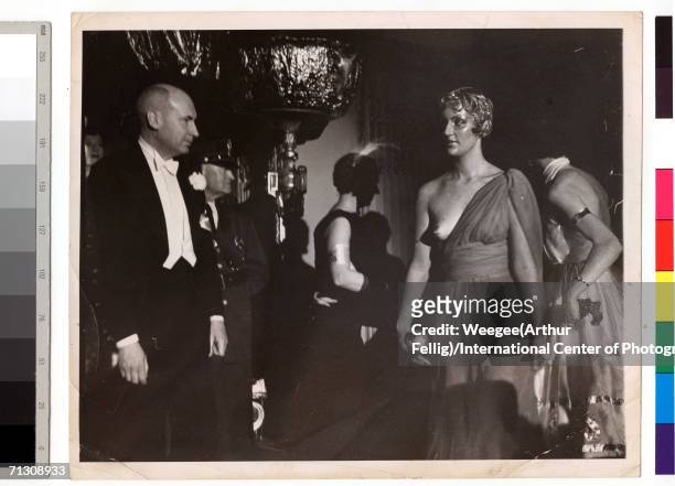 Woman wearing a dress that bares one of her breasts walks past a man wearing coattails at a ball, circa 1945.