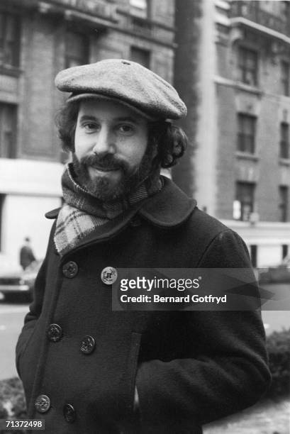 Portrait of American singer and musician Paul Simon as he stands on a sidewalk, hands his the pockets of his pea coat and a cap on his head, 1970s.