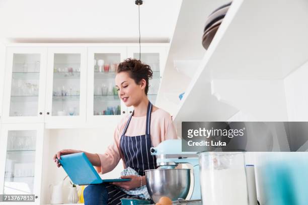 young female baker sitting on kitchen counter looking at laptop - woman baking stock pictures, royalty-free photos & images