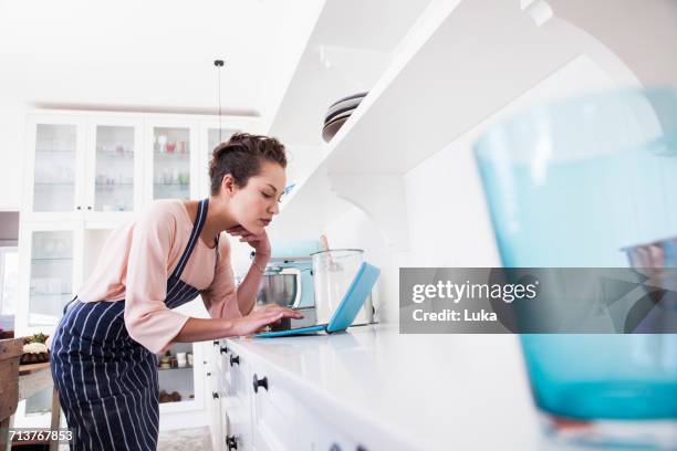 young female baker leaning on kitchen counter looking at laptop - woman baking stock pictures, royalty-free photos & images