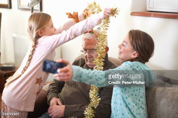 sisters taking smartphone selfie while putting tinsel on sleeping grandfather - christmas humor stock-fotos und bilder