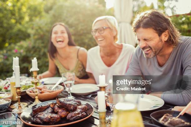 cheerful couple and female friend laughing on dining table during garden party in back yard - friends dinner party stockfoto's en -beelden