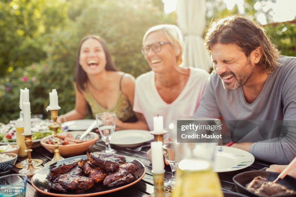 Cheerful couple and female friend laughing on dining table during garden party in back yard