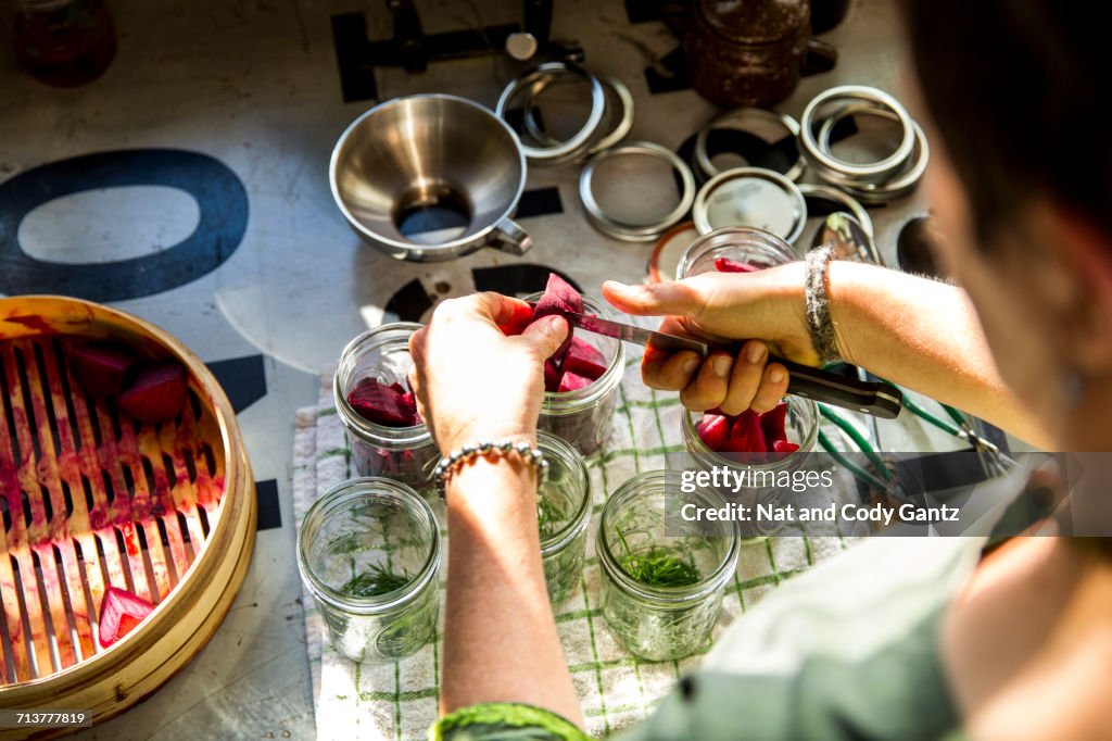 Over the shoulder view of woman slicing beetroot into preserves jar in kitchen