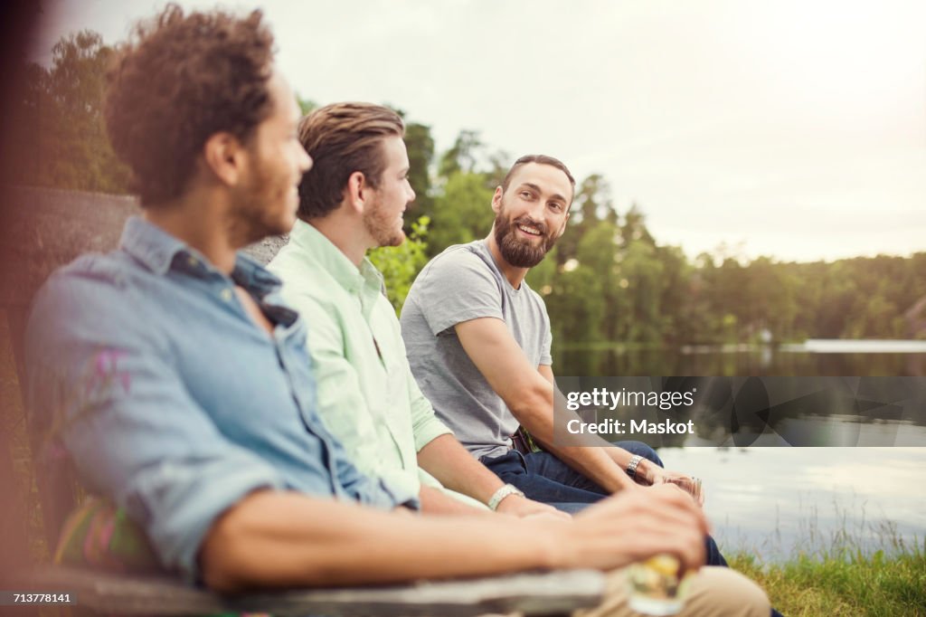 Happy male friends talking while sitting on wooden bench at lakeshore