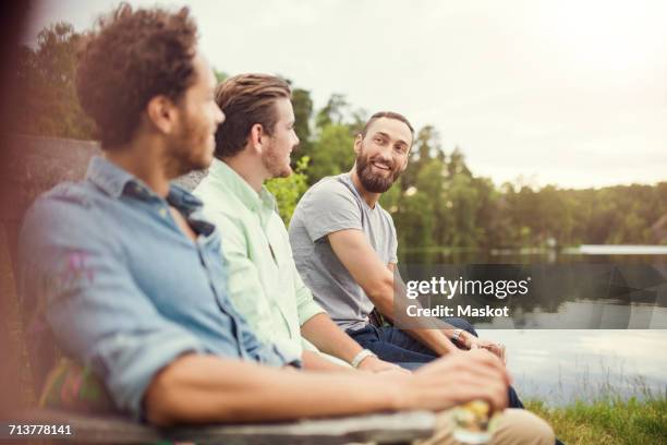 happy male friends talking while sitting on wooden bench at lakeshore - three people sitting stock pictures, royalty-free photos & images