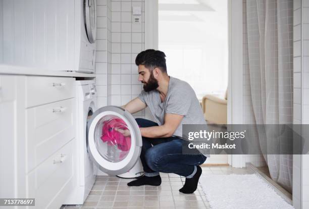 side view of man crouching while loading clothes in washing machine at home - dryer stock-fotos und bilder