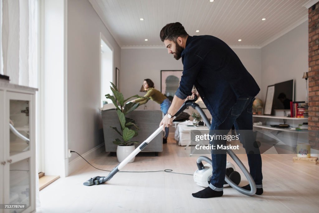 Man vacuuming floor while woman working in living room at home