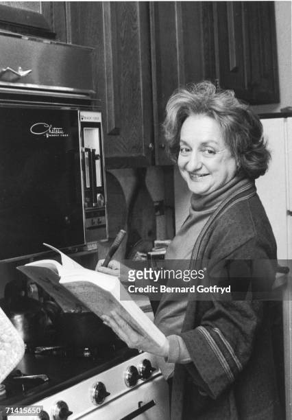 American social activist and writer Betty Friedan turns to look at the camera as she stirs a pot on the stove and holds open a cookbook in a kitchen,...
