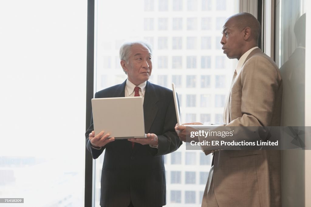 Two businessmen with laptops next to window