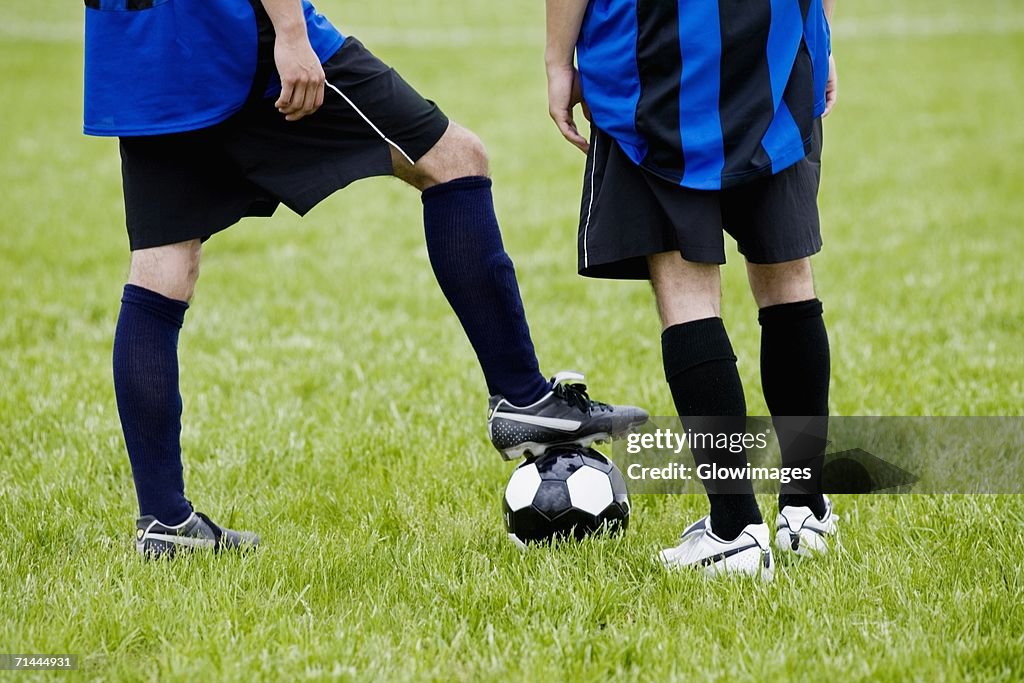 Low section view of a soccer player resting his foot on a soccer ball with another player standing beside him