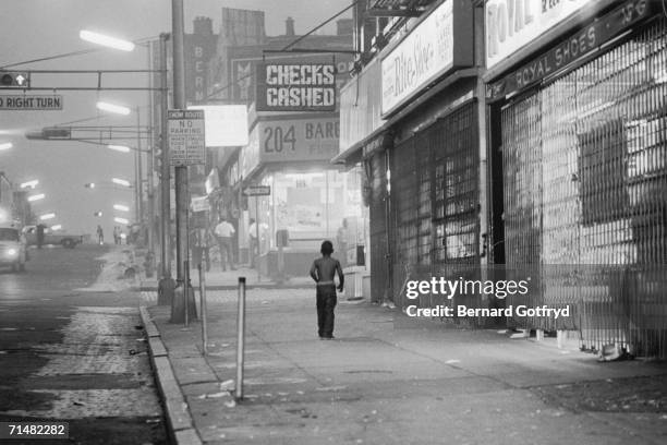 Young, shirtless boy walks down the sidewalk past shuttered storefronts towards a well-lit furniture store around which several men are gathered, in...