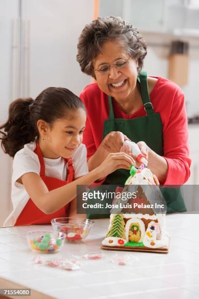 hispanic grandmother helping granddaughter decorate gingerbread house - christmas cake foto e immagini stock