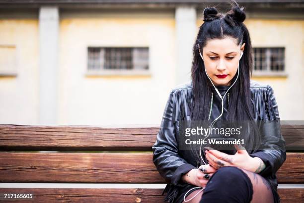 dark-haired young woman sitting on bench listening music with earphones and smartphone - svart rock bildbanksfoton och bilder