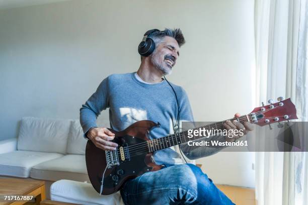 mature man at home playing electric guitar and wearing headphones - freizeitaktivität stock-fotos und bilder